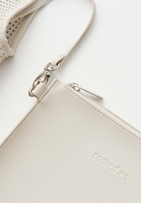 Close-up van een beige leren polstasje met een zilveren rits, metalen sluiting en het geperste woord "someday" op een glad oppervlak.