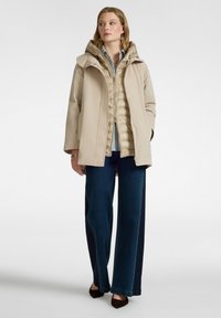 Donna con una giacca beige con cappuccio sopra un gilet trapuntato marrone chiaro, una maglietta a righe, jeans a palazzo bicolore blu e scarpe nere appuntite.