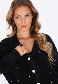 Black, fuzzy cardigan with oversized white buttons and scattered rhinestone accents. Model wearing a ring and large hoop earrings.