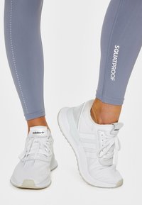 Jasnoszare legginsy z nadrukiem "SQUATPROOF" i kropkowanym wzorem, połączone z białymi sneakersami Adidas z trzema paskami i teksturowaną cholewką.