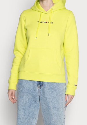 Persoon draagt een felgele Tommy Jeans hoodie met voorvak en lichtblauwe spijkerbroek tegen een effen achtergrond.