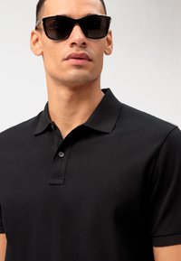 Polo nero con colletto e chiusura a due bottoni, abbinato a occhiali da sole scuri con una montatura classica scura e linee eleganti.