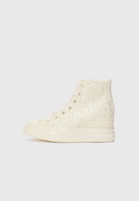 Kremfargede høyhælte sneakers laget av blonder med et teksturert blomster mønster, med gummisåle og flate lisser.