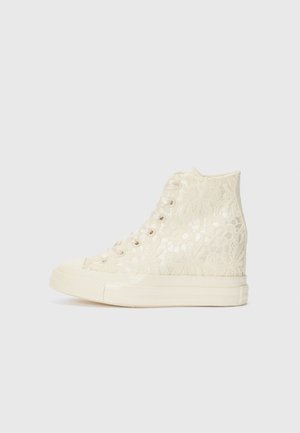 Cream-colored high-top sneaker made of lace with a textured floral pattern, featuring a rubber sole and flat laces.