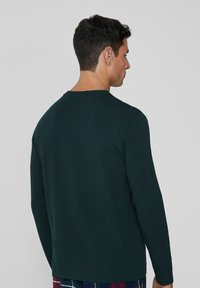 Ciemnozielona koszula z długim rękawem o okrągłym dekolcie i gładkiej fakturze materiału, widoczna od tyłu, zestawiona z kratkowymi spodniami do piżamy.