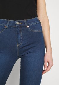 Jeans de mezclilla azul oscuro con un diseño ajustado, que presentan costuras amarillas, un botón plateado y bolsillos frontales. Textura suave y corte moderno.