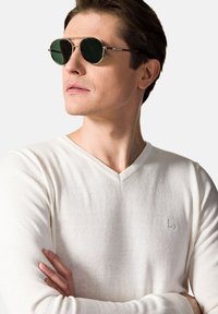 Okrągłe okulary przeciwsłoneczne z zielonymi soczewkami i złotą, metalową oprawką, w zestawieniu z białym swetrem z dekoltem w serek z subtelnym logo marki.