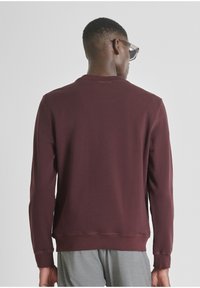 Sudadera burdeos de mangas largas, con cuello redondeado y dobladillo acanalado. Presenta un minimalista detalle de logo en la parte posterior. Textura suave.