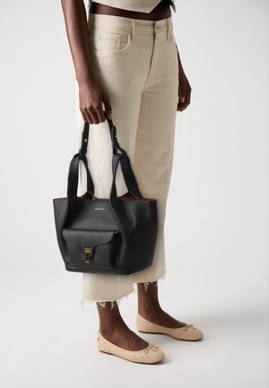 Black leather tote bag with a textured finish, gold hardware, and front pocket; held by a person wearing light beige pants and ballet flats.