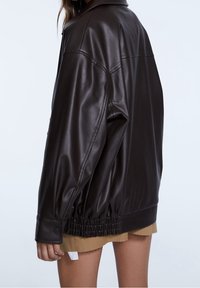 Brązowa kurtka z sztucznej skóry o luźnym kroju, z elastycznym dołem i mankietami na guziki. Posiada klasyczny kołnierz i gładką teksturę.