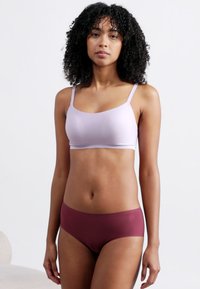 Γυναίκα με σγουρά μαλλιά, φορώντας ένα ανοιχτό μοβ σουτιέν και μπορντό εσώρουχα, στέκεται μπροστά σε ένα απλό λευκό φόντο.