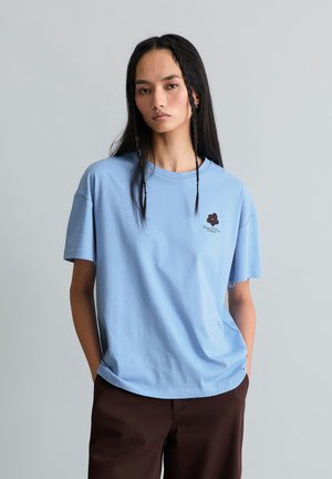 Frau mit langen schwarzen Haaren, die ein hellblaues T-Shirt mit kleinem dunklem Blumenlogo und dunkelbraune Hose trägt, vor einem grauen Hintergrund stehend.