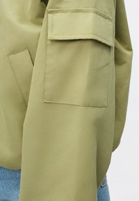 Olivenfarget jakke med ermer laget av glatt stoff, med en firkantet lomme og rette sømmer. Blå denim under vises.