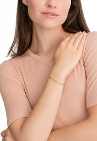 Die goldene Kettenarmband hat verlängerte Glieder. Das Model trägt ein hellrosa, eng anliegendes Shirt, mit einem Arm, der über ihrer Brust ruht.