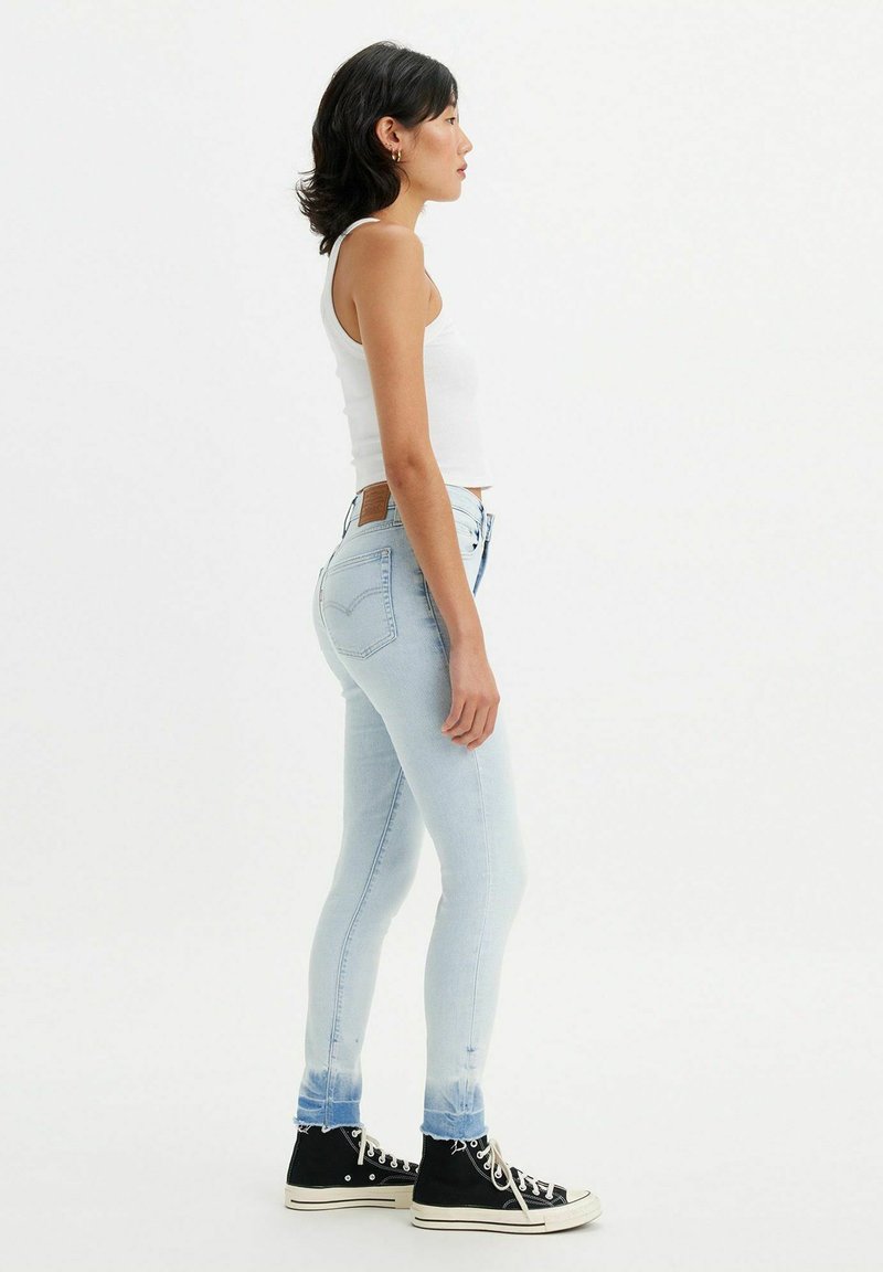 Dünne, hellblaue Jeans mit einem ausgewaschenen Saum, getragen mit schwarzen High-Top-Sneakern. Verfügt über ein hellbraunes Lederpatch am hinteren Bund.