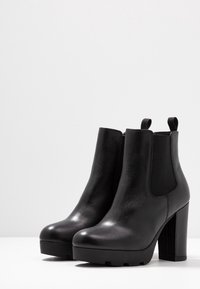 Une paire de bottines en cuir noir avec des talons épais et des panneaux latéraux en élastique sur une surface blanche.
