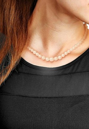 Collier de perles rose clair avec des tailles graduées, fabriqué dans un matériau lisse, porté contre un haut noir, reposant sur la clavicule.