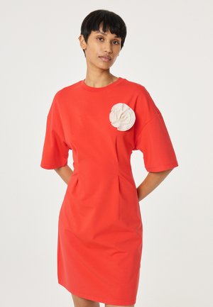 Junge Person trägt ein knallrotes, knielanges Kleid mit kurzen Ärmeln und einer großen weißen Stoffblume nahe der Schulter.