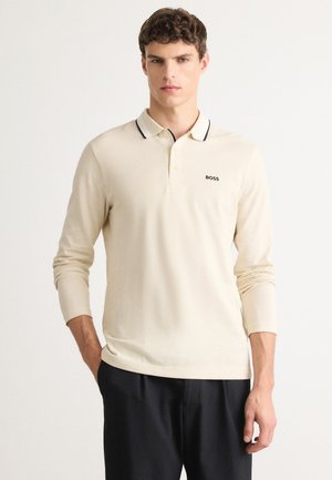 Ung mand iført en cremefarvet langærmet polo med sort trim og "BOSS" logo, parret med sorte bukser, stående foran en ensfarvet baggrund.