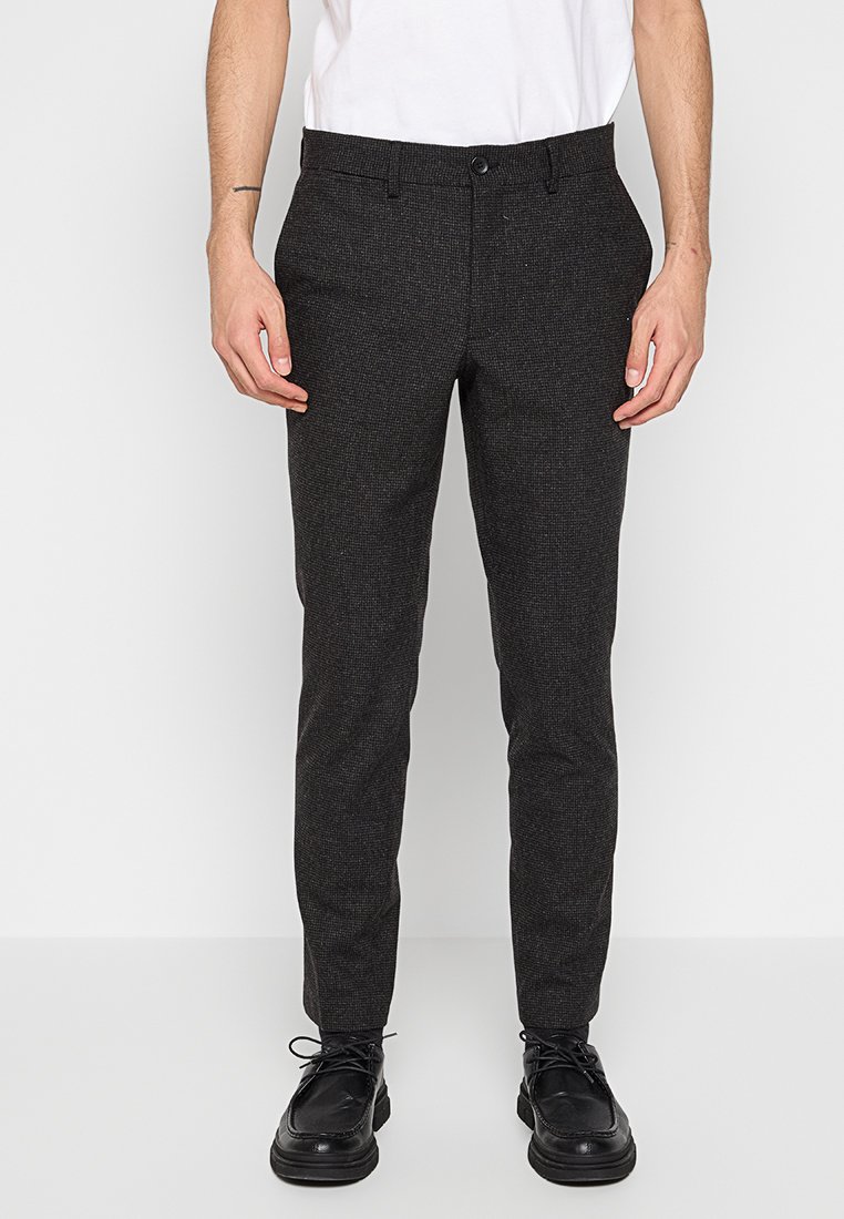 Dunkelgraue maßgeschneiderte Hosen mit einem subtilen strukturierten Muster, Gürtelschlaufen, Fronttaschen und einem Knopfverschluss, kombiniert mit schwarzen Schuhen.