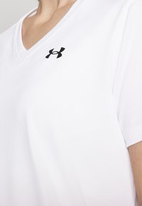 Valkoinen urheilupaita, jossa on V-pääntie, lyhyet hihat ja pieni musta Under Armour -logo vasemmassa rintamuksessa. Silkkinen rakenne.