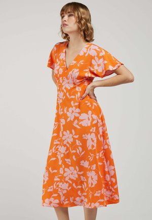 Femme portant une robe midi orange à imprimé floral rose, manches courtes, col en V, debout avec une main sur la hanche devant un fond uni.