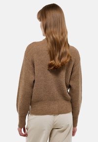Femme aux longs cheveux roux portant un pull en maille marron et un pantalon beige clair, debout de dos sur un fond uni.