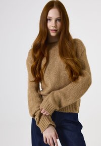 Strickpullover in Hellbraun, mit lockerer Passform, breiten gerippten Bündchen und strukturiertem Stoff. Kombiniert mit dunkelblauen Jeans.