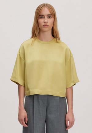 Korte mouw, cropped top in zachte, gladde gele stof met een ronde halslijn en een ontspannen pasvorm. Gedragen met grijze, geplooide broeken.