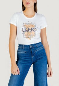 Camiseta blanca con un diseño gráfico floral y texto "LIU J" en azul oscuro. Combinada con vaqueros azules de cintura alta.