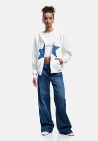 Weiße Zip-Hoodie mit blauen Sternen-Patches, kombiniert mit einem cropped weißen Top und lockeren blauen Jeans. Hat weite Beine und einen entspannten Schnitt.