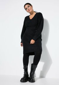 Schwarzes, glitzerndes Strickkleid mit V-Ausschnitt und langen Ärmeln, kombiniert mit schwarzen Leggings und klobigen schwarzen Stiefeln. Schlichtes, elegantes Design.