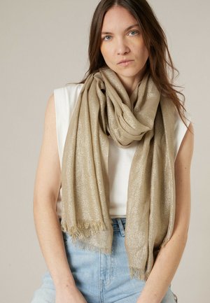 Femme aux longs cheveux bruns portant une écharpe beige brillante sur un débardeur blanc et un jean bleu clair sur un fond uni.