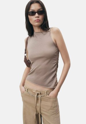 Top senza maniche color beige con arricciatura laterale, abbinato a pantaloni beige chiaro con una cintura in catena argentata. La modella indossa occhiali da sole scuri.
