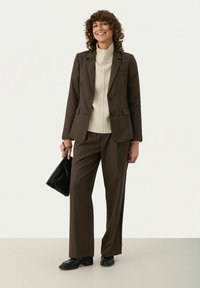 Bruin op maat gemaakt pak met wijde broekspijpen en een bijpassende blazer. Crème geribde coltrui eronder. Zwart leren handtas en hakken.