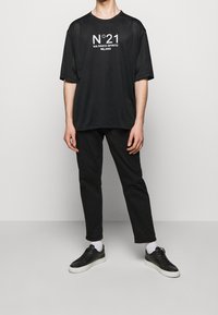 Černé síťované tričko s krátkým rukávem s bílým nápisem "N°21 VIA SANTO SPIRITO MILANO," v kombinaci s černými kalhotami a černými teniskami.