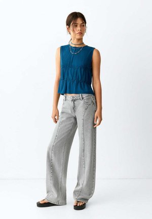Femme portant un haut bleu sans manches, un jean gris clair à jambes larges, des sandales à plateforme noires et des colliers superposés, debout devant un fond blanc uni.