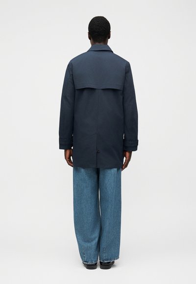 Ναυτικό παλτό με πίσω γιάκα, μακριά μανίκια και ίσια γραμμή, συνδυασμένο με ανοιχτό μπλε τζιν παντελόνι φαρδιάς γραμμής και μαύρα παπούτσια.