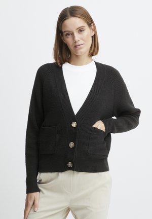 Black knitted cardigan with a V-neck, featuring four large buttons and two front pockets. Ribbed texture and relaxed fit, paired with light trousers.
