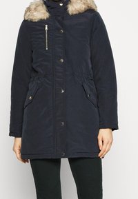 Navy blue parka jacket with faux fur hood, zippered pocket, snap closure, elastic waist, and side pockets. Smooth textured fabric.