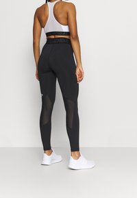 Leggings atletici neri con pannelli in rete, vita alta con logo, abbinati a una maglietta bianca con schiena scoperta e scarpe da ginnastica bianche.