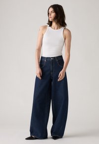 Jean en denim bleu foncé à jambes larges et taille haute, avec des détails de poche arrière et des coutures contrastées. Associé à un débardeur blanc.