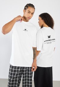 Balta per didelė t-shirt su juodu drugelio logotipu priekyje ir gale. Tekstas gale skamba "METAMORFOZĖ" ir "SKRISK". Kasdienis fasonas.