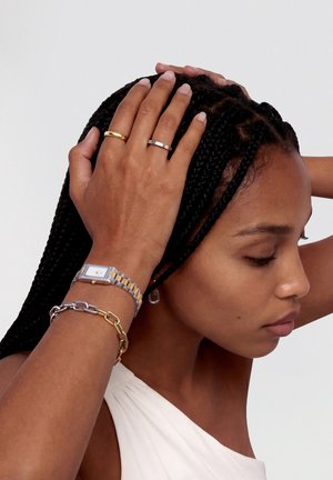 Žena s copánky drží hlavu oběma rukama, na prstech má zlaté a stříbrné prsteny, náramkové hodinky dvoubarevné a náramky z řetízků různých kovů.