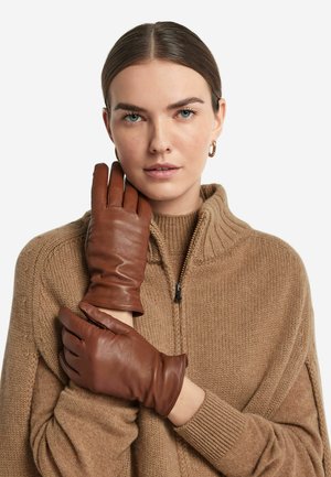 Brune lædersludder med en glat tekstur og tætsiddende design, der har fire fingre og en stram pasform, båret af en model i en beige sweater.