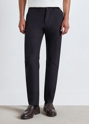 Uomo con pantaloni neri a taglio dritto e mocassini in pelle marrone scuro, in piedi su un pavimento chiaro davanti a uno sfondo semplice.