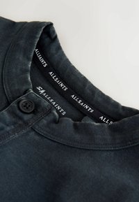 Detailný záber na tmavosivý golier košele s čiernym gombíkom a viditeľnou značkou AllSaints vo vnútri výstrihu.