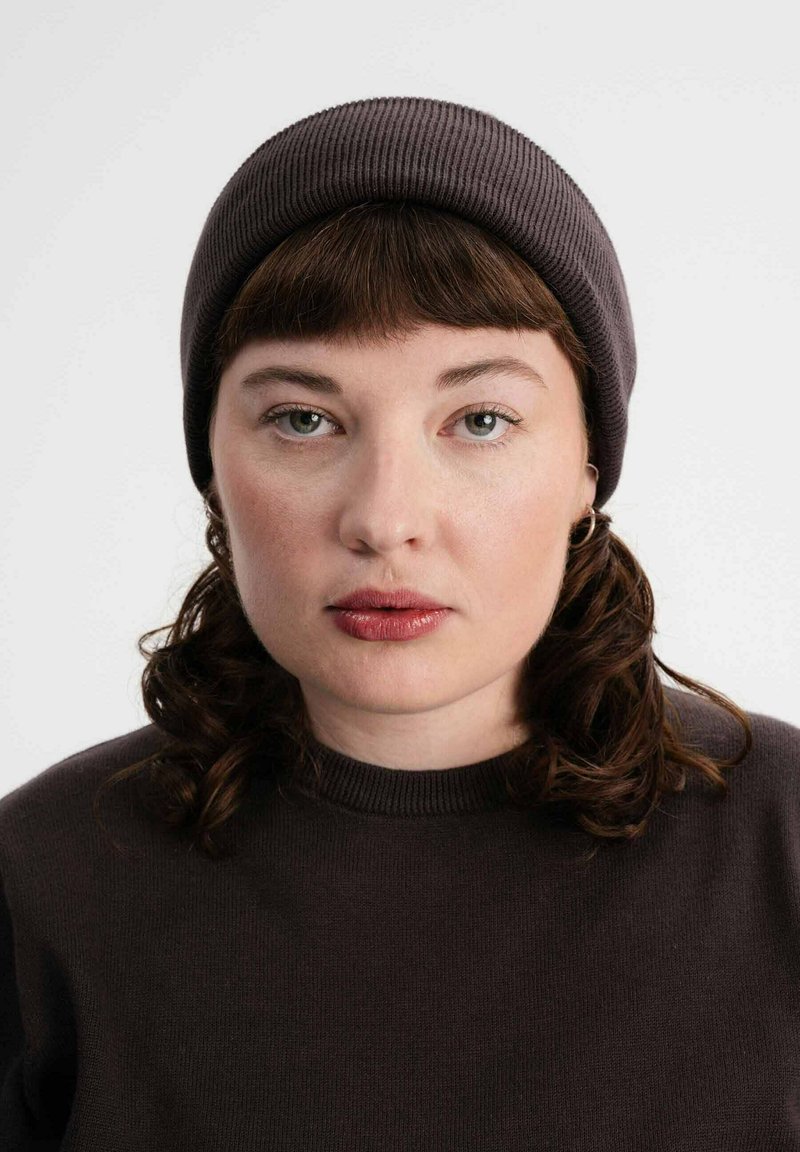 Donkergrijze gebreide beanie op een persoon met schouderlang krullend haar. Ze dragen een bijpassende donker trui met een ronde hals.