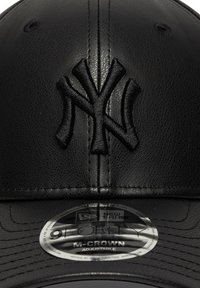 Melna ādas cepure ar iegravētu Ņujorkas "Yankees" logo, regulējama izmēra, un ar apļveida birku, kas atzīmē to kā New Era 9FORTY M-CROWN.