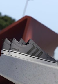 Zapatillas Adidas grises con rayas negras descansando sobre un saliente de piedra, con un fondo difuminado de una estructura geométrica roja y cielo azul.
